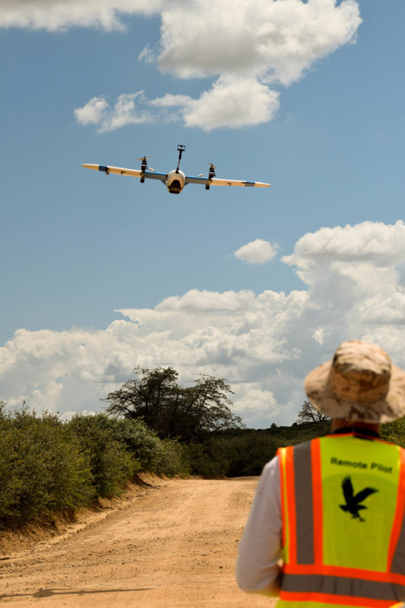 A fixed wing unmanned aerial vehicle flies toward the camera above a dirt road in mostly clear skies, while the remote pilot stands to the side. The UAV has a boom protruding from its nose, which supports a TriSonica Mini Wind and Weather Sensor.