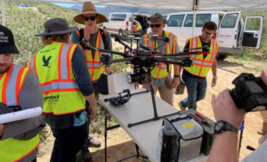 Technicians, standing under a sun shelter in rolling Arizona hills, prepare a multi-rotor UAV, equipped with a LiDAR mapping payload, for flight.